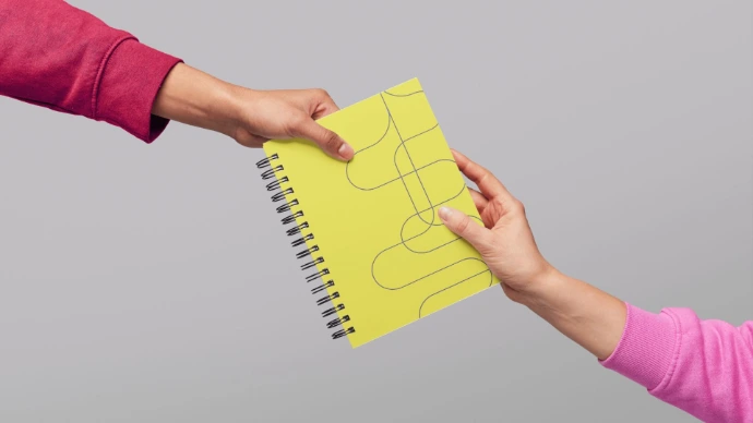 dois braços diferentes, um vestindo uma blusa rosa escuro, outra rosa claro, seguram um caderno juntas.